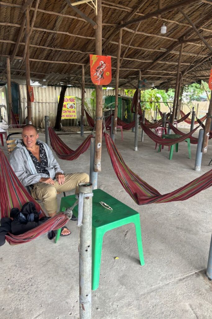 A traveler relaxing in a shaded roadside rest stop filled with hammocks tied between wooden posts, a break point on the journey to Cai Be Village in southern Vietnam.