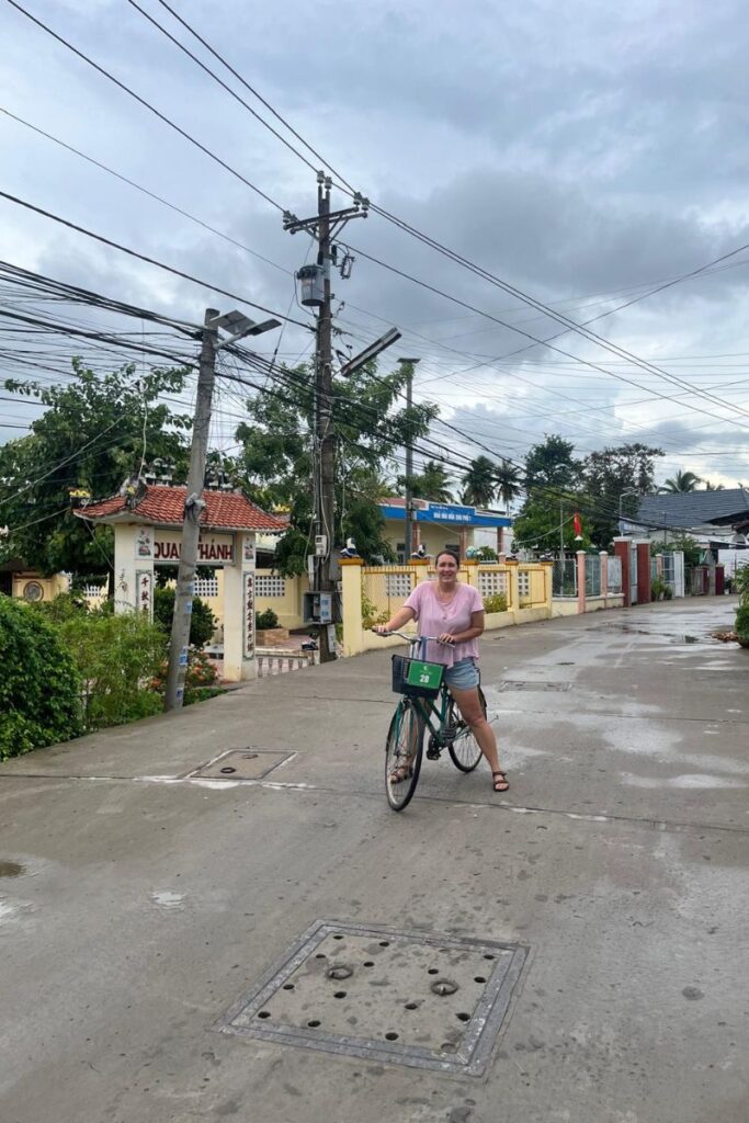 Traveler riding a green rental bicycle through a quiet, residential street in Cai Be Village, Vietnam, with colorful buildings, overhead power lines, and cloudy skies in the background.
