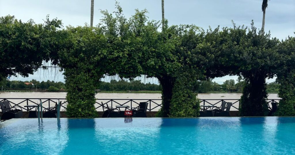 Infinity pool at Mekong Lodge in Cai Be Village, overlooking the Mekong River with lounge chairs shaded by lush green archways and a backdrop of palm trees and blue sky.