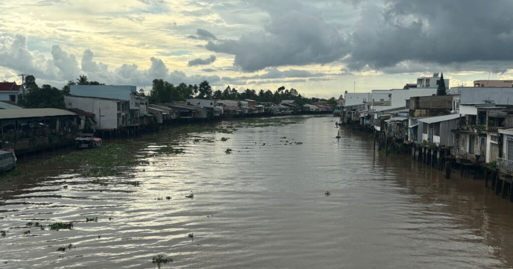 View of the brown river in Cai Be Vietnam Mekong Delta, lined with stilted riverside homes and shops under a moody, cloud-filled sky, with floating vegetation scattered on the water.

