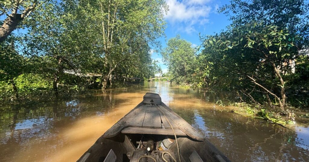 View from inside a wooden canoe gliding through a narrow, muddy canal in Cai Be Vietnam, surrounded by thick green trees on both sides and reflecting a bright blue sky overhead.
