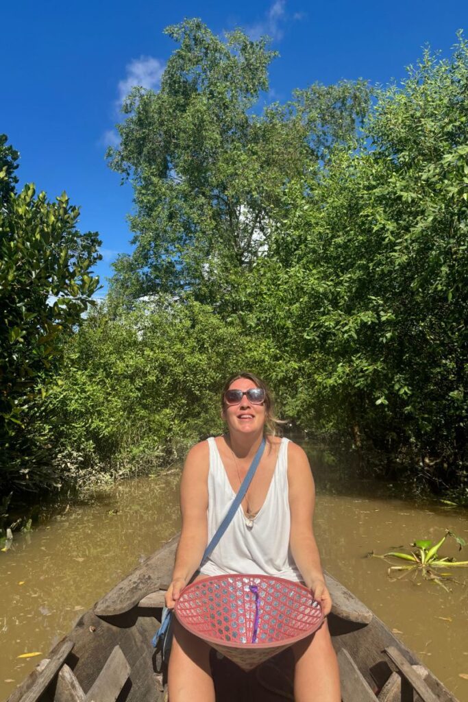 
Traveler smiling in a wooden sampan on a narrow canal in the Cai Be Vietnam Mekong Delta, holding a pink-patterned conical hat and surrounded by dense green vegetation under bright sunlight.