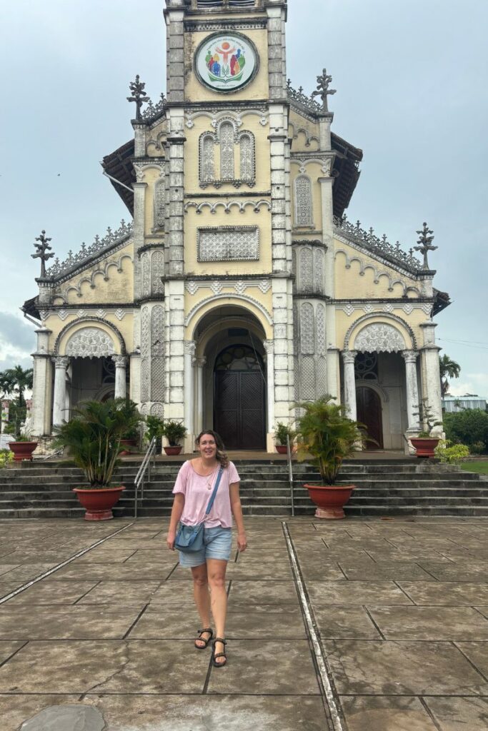 Traveler walking in front of the French colonial–style Cai Be cathedral in Cai Be Vietnam, with large wooden doors, potted plants, and wide stone steps leading up to the entrance.