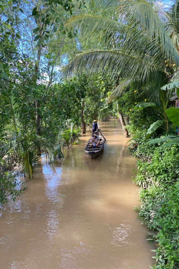 A wooden sampan glides through a narrow, muddy canal in Cai Be, surrounded by lush tropical greenery. A local guide wearing a conical hat steers from the back while two passengers sit at the front, enjoying the quiet, shaded route through the Mekong Delta.