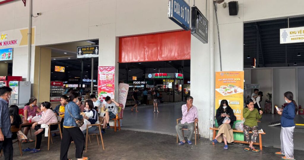 Travelers sitting and standing at a rest stop halfway between Ho Chi Minh City and Can Tho, with seating areas, food vendors, and signage under a covered building.