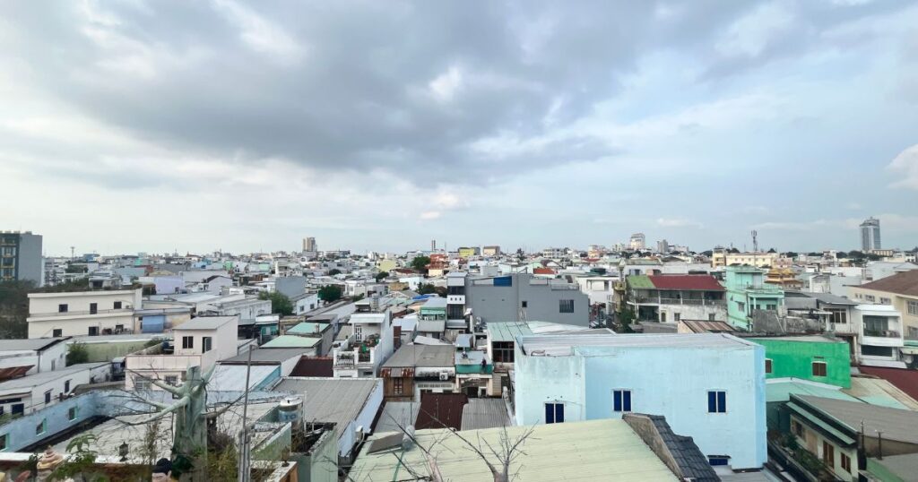 Panoramic view over Can Tho city from a rooftop, showing a dense cluster of colorful low-rise buildings under a cloudy afternoon sky.