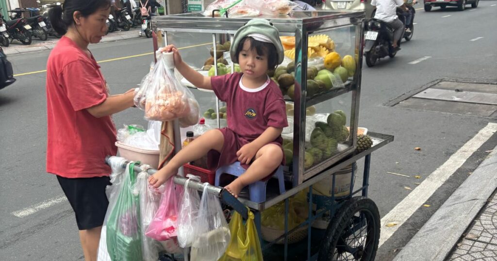 A young girl sits on a small stool next to a street fruit cart in Ho Chi Minh City.