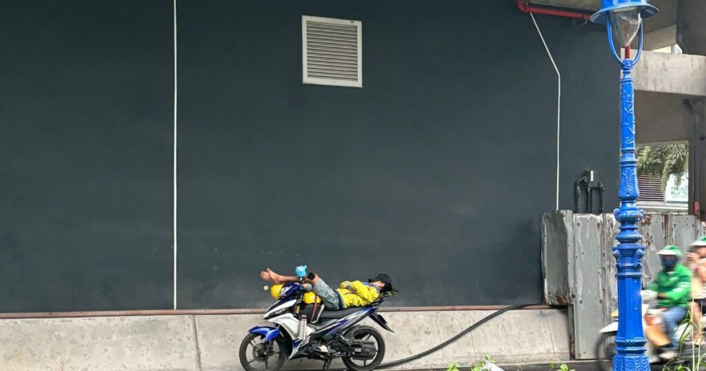 A man naps stretched out across a motorbike under a bridge in Ho Chi Minh City, a reminder that not all street scenes are tied to Vietnamese scams.