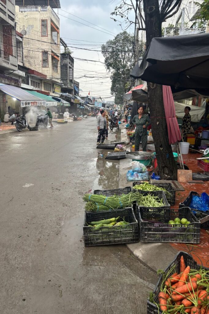 Vendors selling fresh vegetables and seafood at a wet street market near the wharf in downtown Cantho, with locals shopping under tarps and motorcycles passing through the narrow lane.