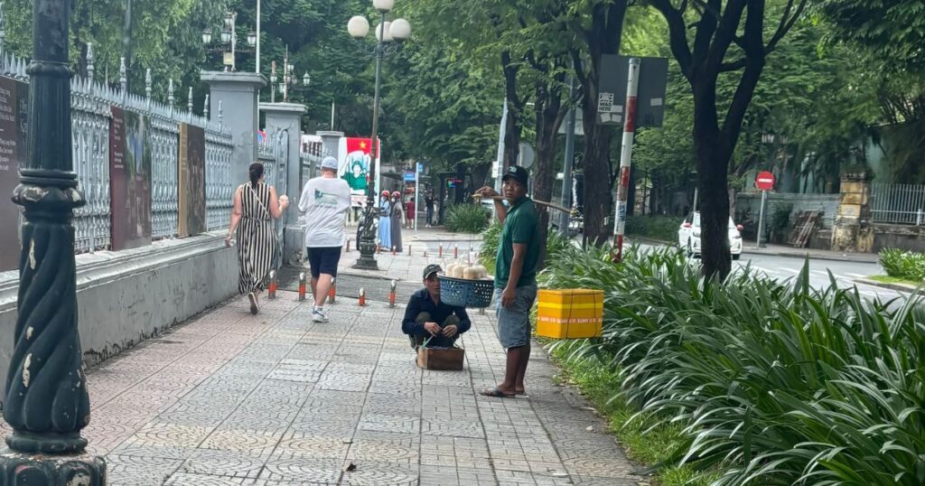 Two coconut sellers approach tourists on a sidewalk in Ho Chi Minh City, a common scene linked to scams in Vietnam.