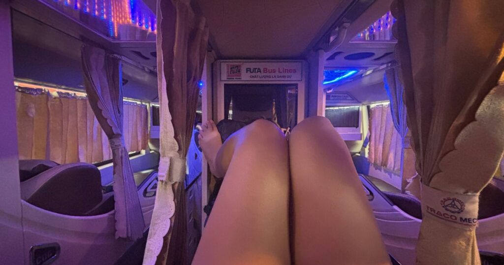 Passenger’s legs stretched out on a sleeper bus from Saigon to Can Tho, with purple LED lighting, privacy curtains, and the Futa Bus Lines logo visible inside the vehicle.