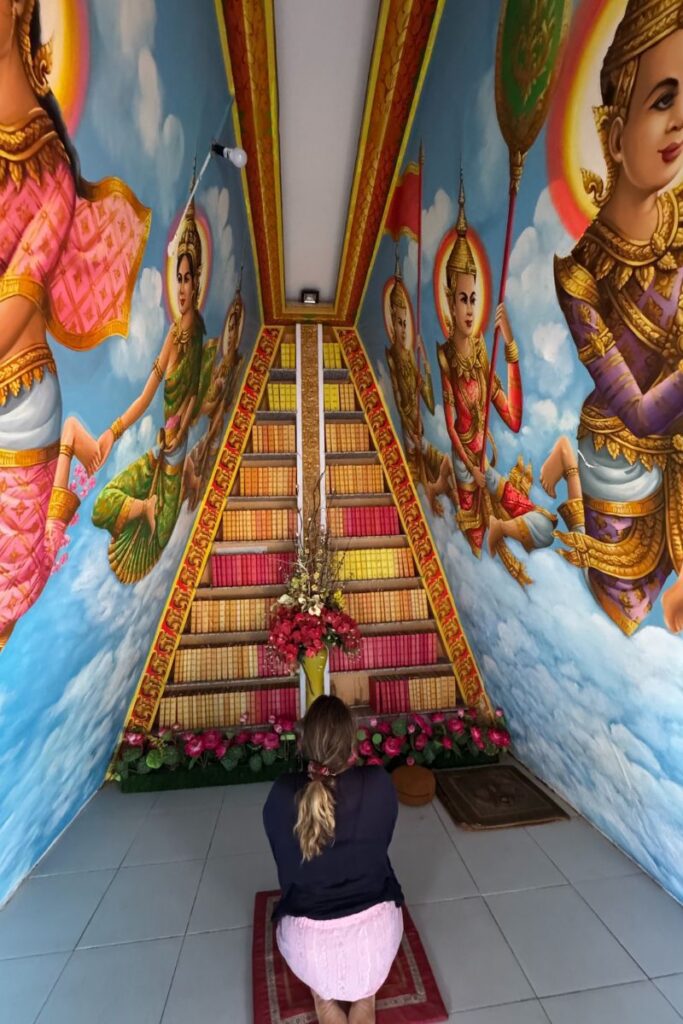 Traveler kneeling in front of a colorful book shrine at a Buddhist pagoda in Cantho, surrounded by painted figures, clouds, and a staircase-like display of stacked scriptures and offerings.