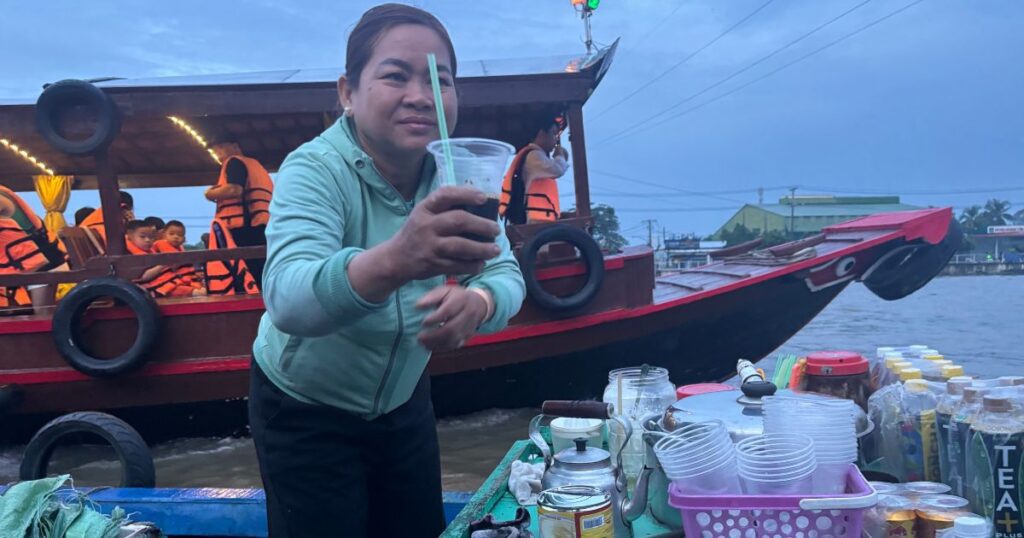 A woman serving iced coffee from her boat at the Can Tho floating market during early morning hours, with plastic cups and bottled drinks arranged in front and other passengers in life vests on a nearby boat.
