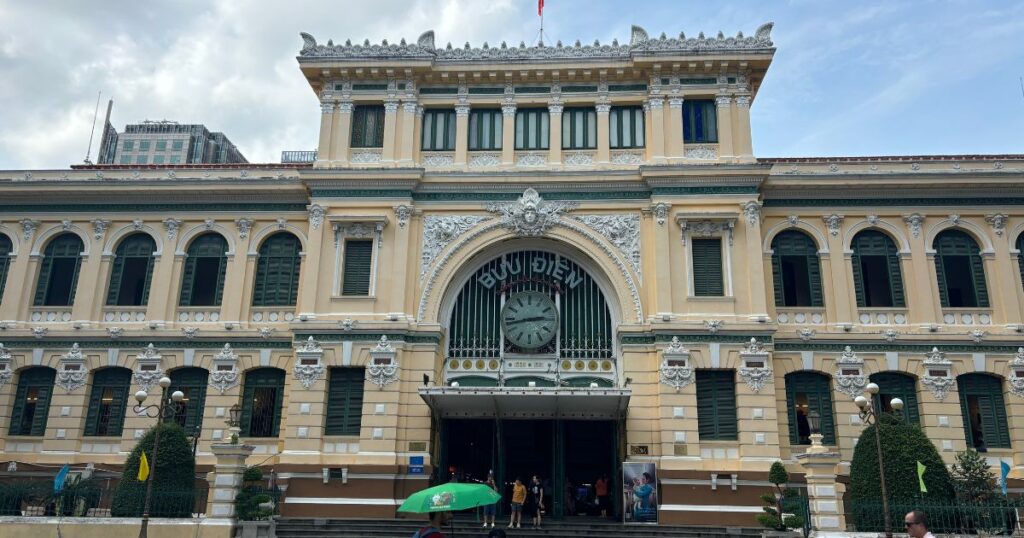Outside view of the Central Post Office in Ho Chi Minh City with pale yellow walls, green shutters, a large arched entrance, and a bright blue sky, a popular HCMC attraction.