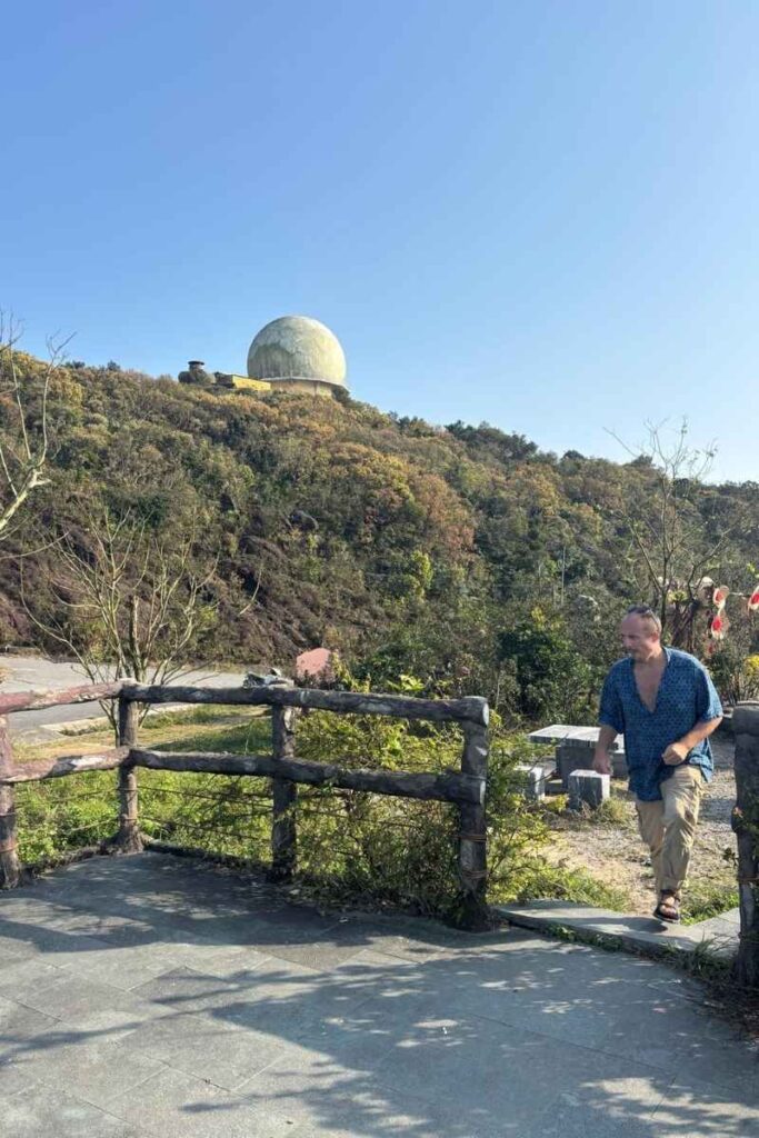 A man walks along a scenic viewpoint atop Monkey Mountain Da Nang, with a large radar dome perched on a nearby hill and surrounded by autumn-toned foliage under a clear blue sky.