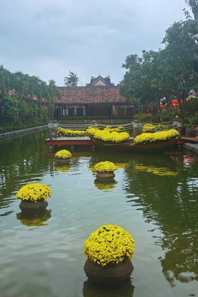 Entrance area of Ba Na Hills with a pond, floating yellow flowers, traditional-style buildings, and decorative boats near the ticketing zone.