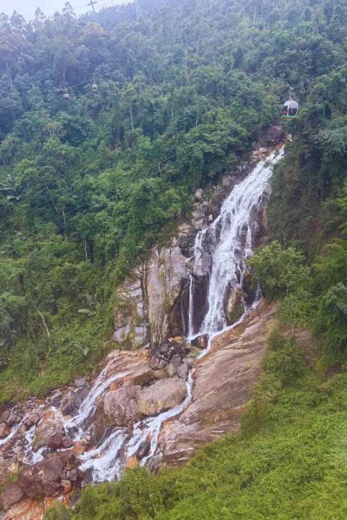 View from a Ba Na Hills cable car passing over a tall waterfall surrounded by dense green forest, heading toward the Golden Bridge Ba Na Hills area.
