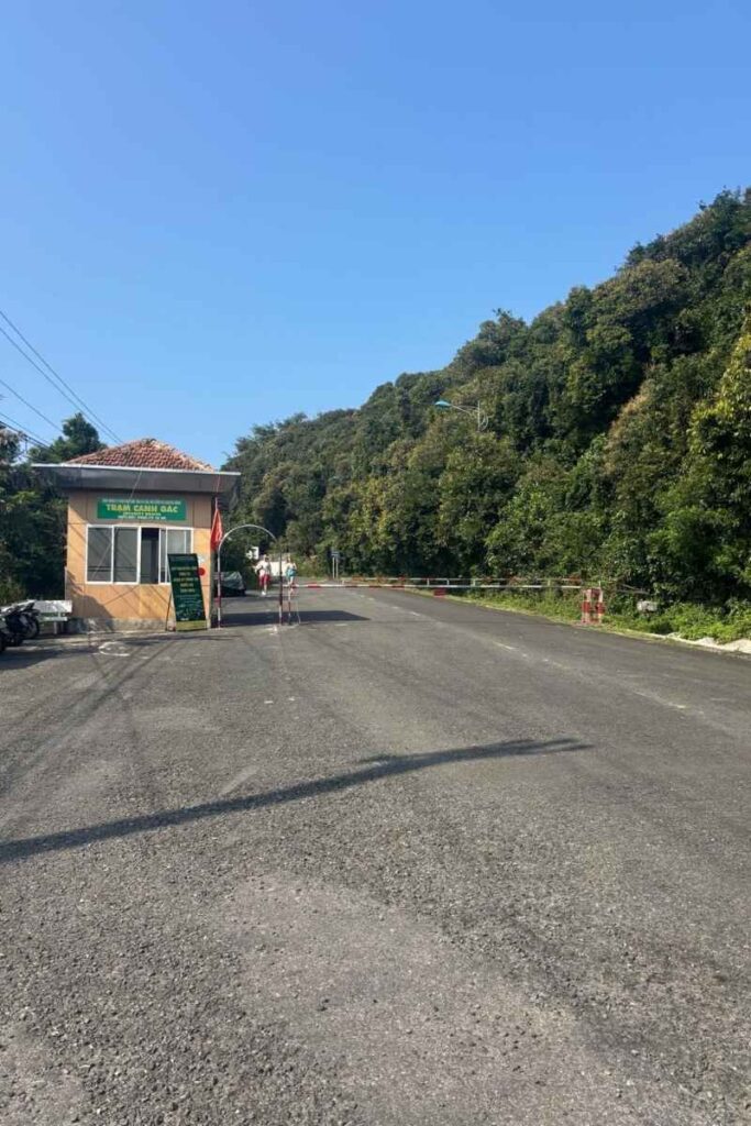 A roadside checkpoint at on the road up Monkey Mountain Da Nang, with a small tan guard booth, a green sign in Vietnamese, and a barrier gate leading into a forested mountain road under a clear blue sky.
