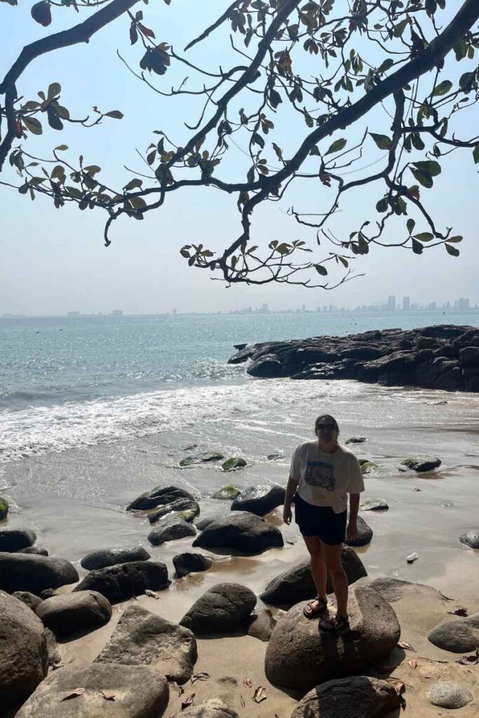 A woman stands on a large rock at a secluded beach near Monkey Mountain Da Nang, with gentle waves washing over the shore, smooth boulders scattered along the sand, and city buildings visible across the bay in the background.