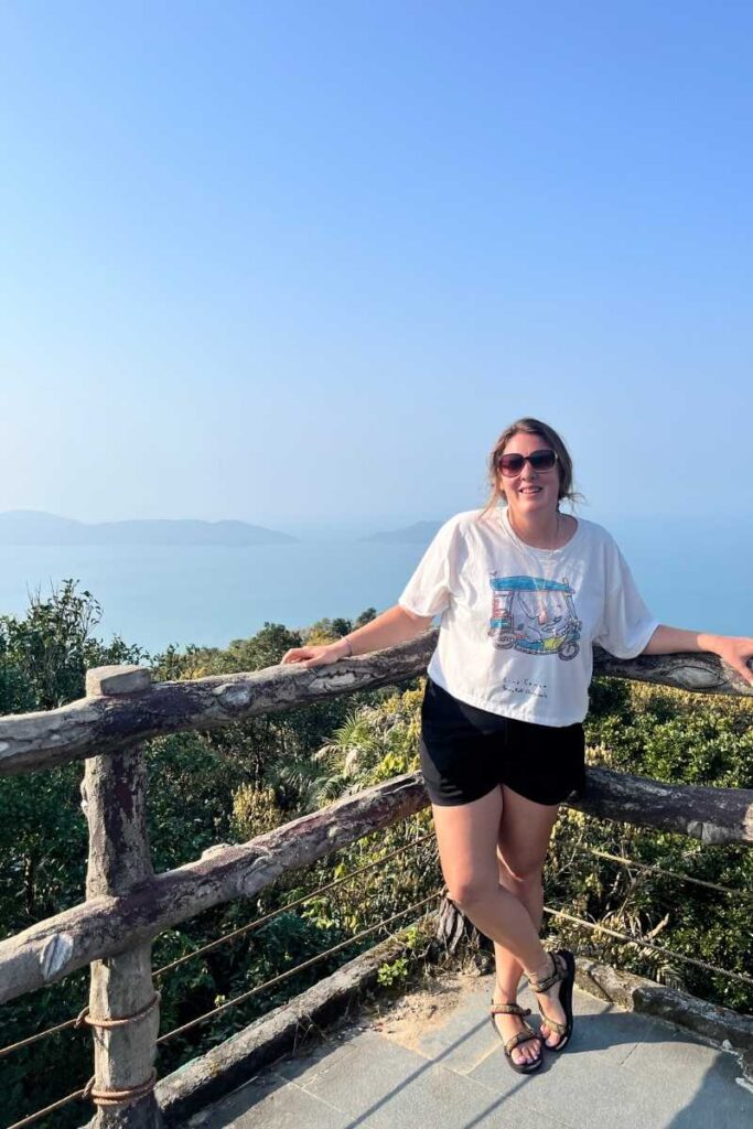 A smiling woman poses at a scenic wooden lookout on Monkey Mountain Da Nang, with forested hills and the hazy blue sea stretching into the distance under a clear sky.