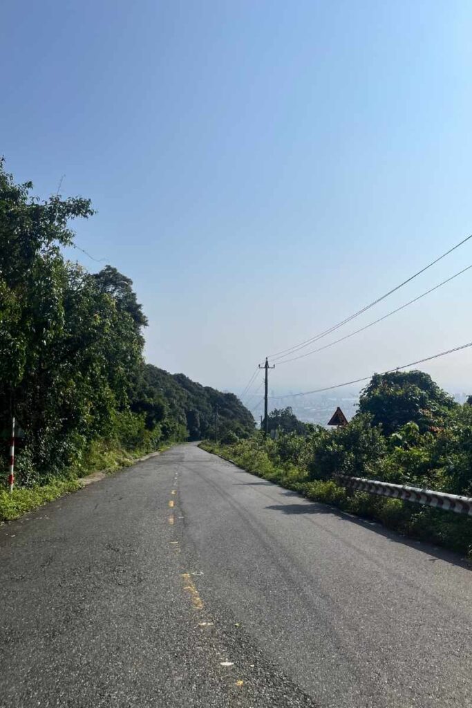 A winding road cutting through dense greenery on Monkey Mountain Da Nang, with power lines overhead and a distant view of Da Nang city barely visible through the hazy backdrop.