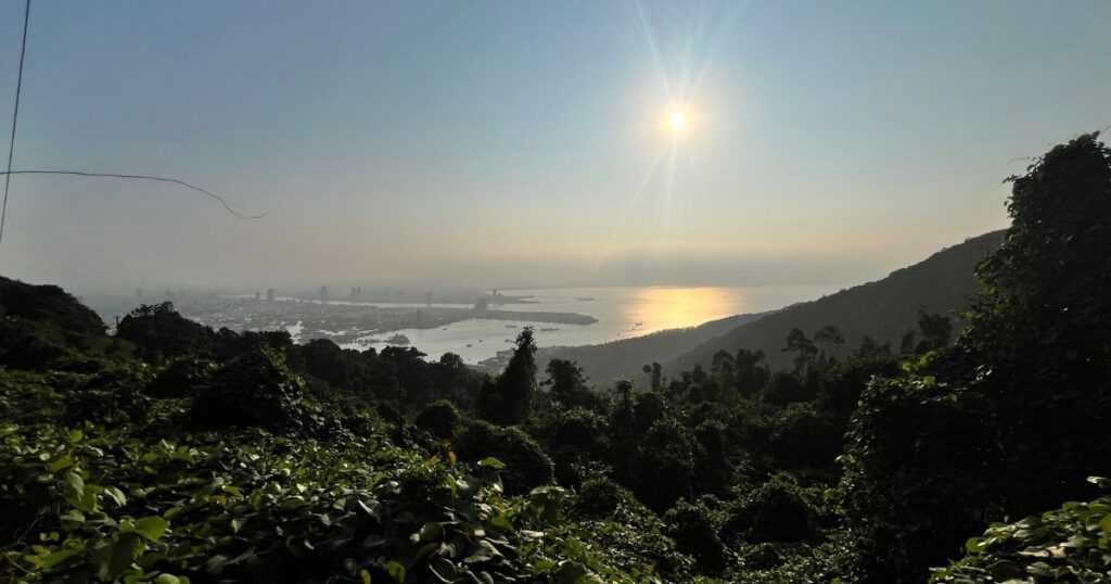 A breathtaking sunset view from Monkey Mountain Da Nang, with the sun casting a golden reflection over the sea, and the lush green forest below framing the distant cityscape of Da Nang.