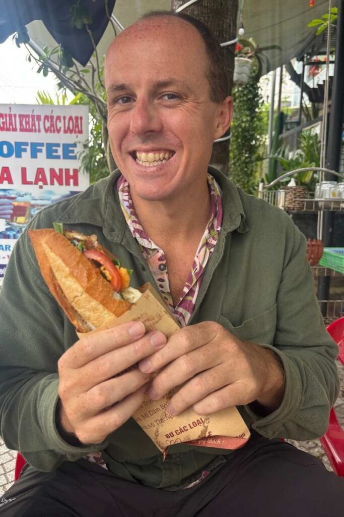 Person smiling while holding a large bánh mì sandwich at a food stall in Da Nang.