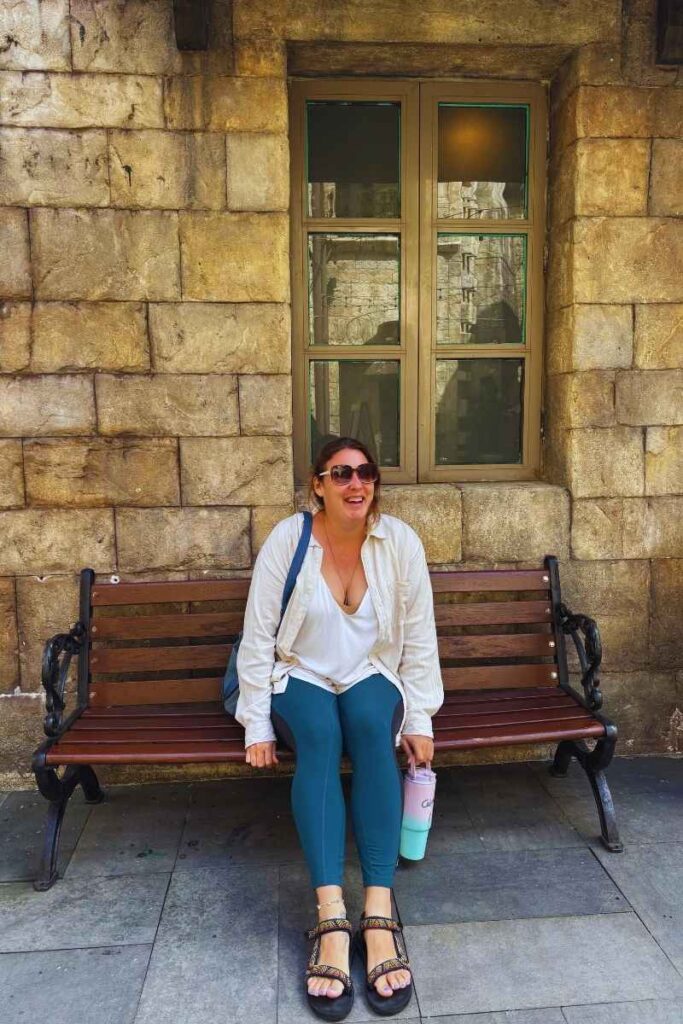 Woman sitting on a wooden bench in front of a stone wall at Ba Na Hills, wearing sunglasses and holding a tumbler.