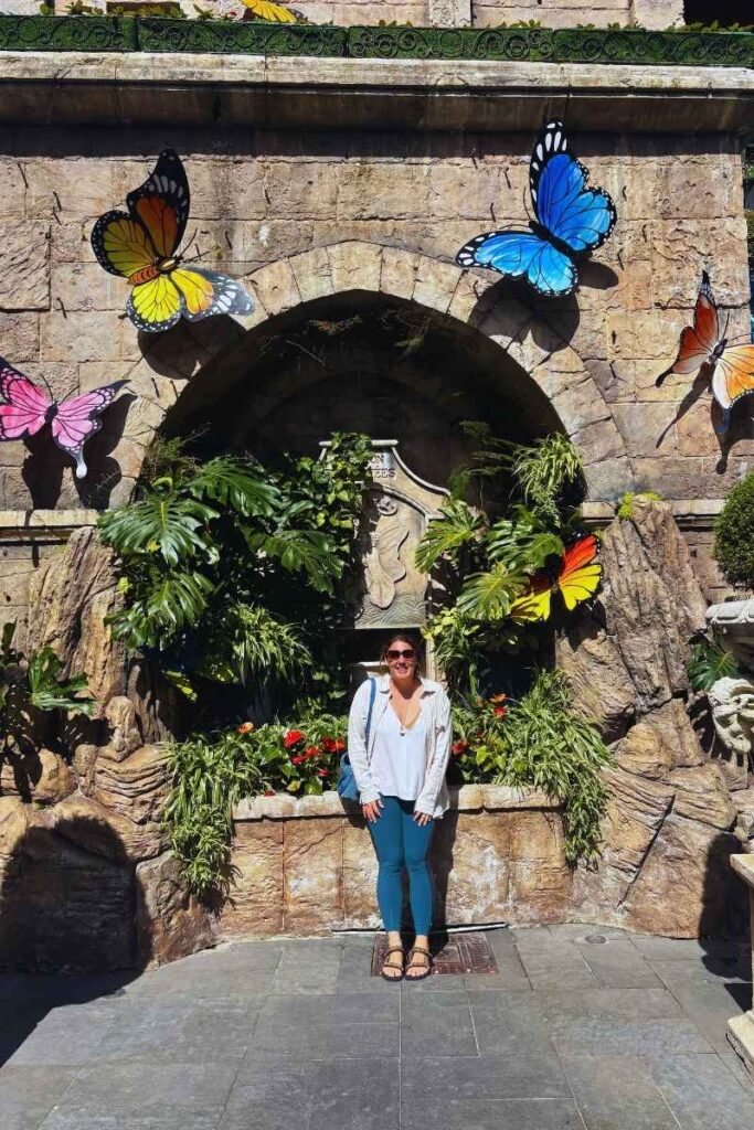 Visitor posing in front of a decorative stone fountain at Ba Na Hills, surrounded by tropical plants, colorful butterfly decorations, and a carved plaque.