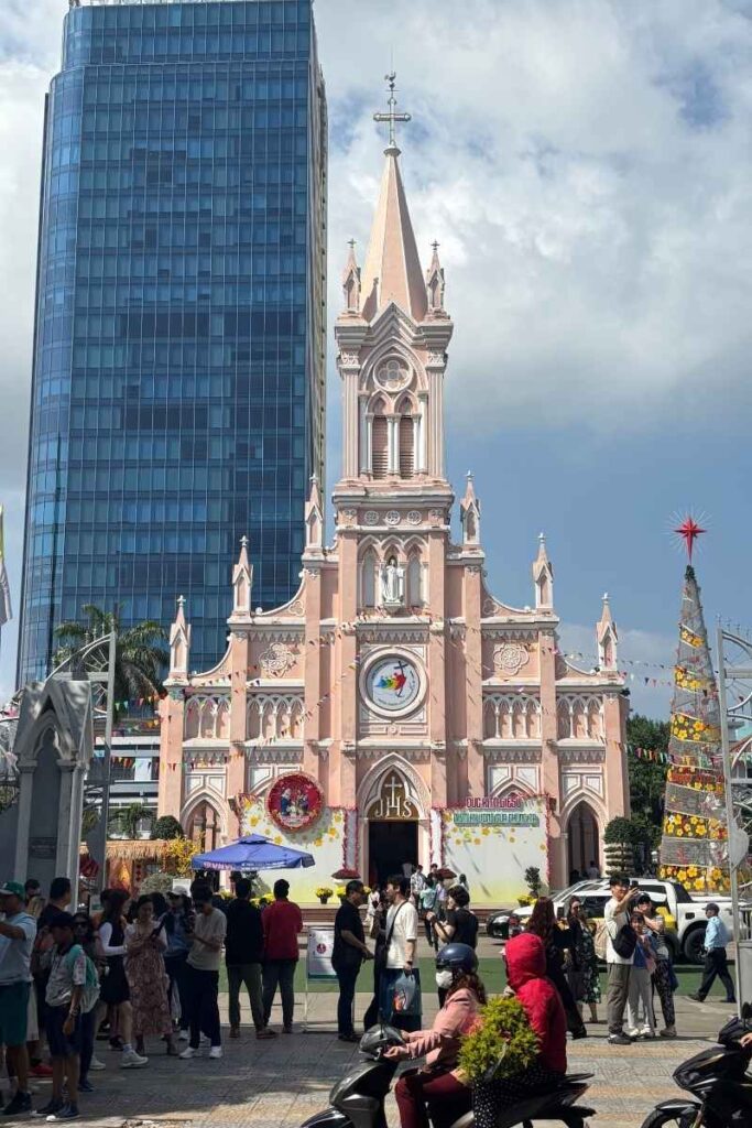 The pink Da Nang Cathedral with people in front, set against a blue sky and modern buildings.