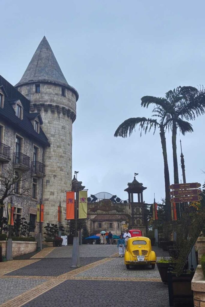 Stone-paved street at Ba Na Hills French Village with a vintage yellow car parked near a signpost, castle-style buildings, and tourists walking toward an archway.