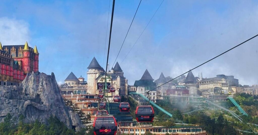 Cable cars arriving at the mountaintop French Village area of Ba Na Hills, with castle-like buildings, colorful towers, and stone structures visible under a clear blue sky.
