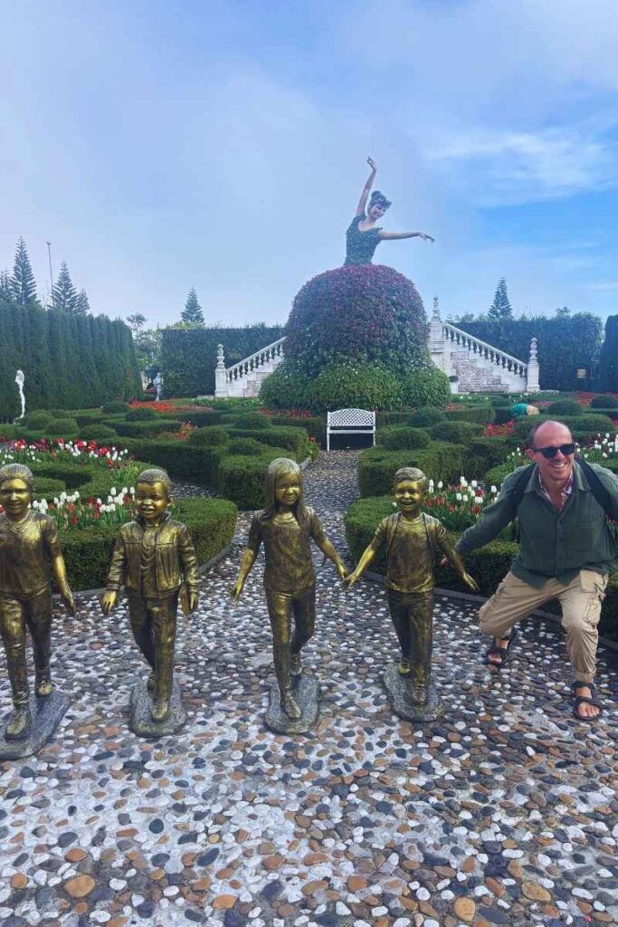 Man posing with a group of bronze statues of children holding hands in the gardens near the Ba Na Hills Gardens.