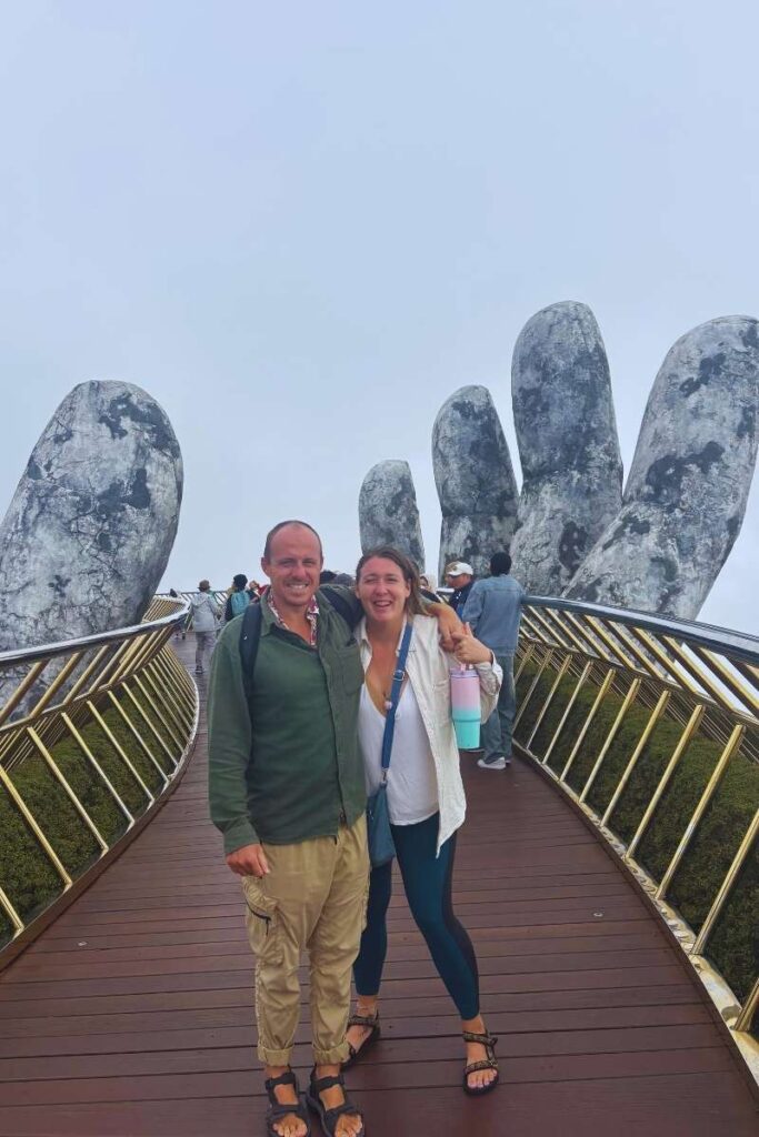 Two people posing on the Golden Bridge at Ba Na Hills, with the famous giant stone hands holding the bridge in the background.