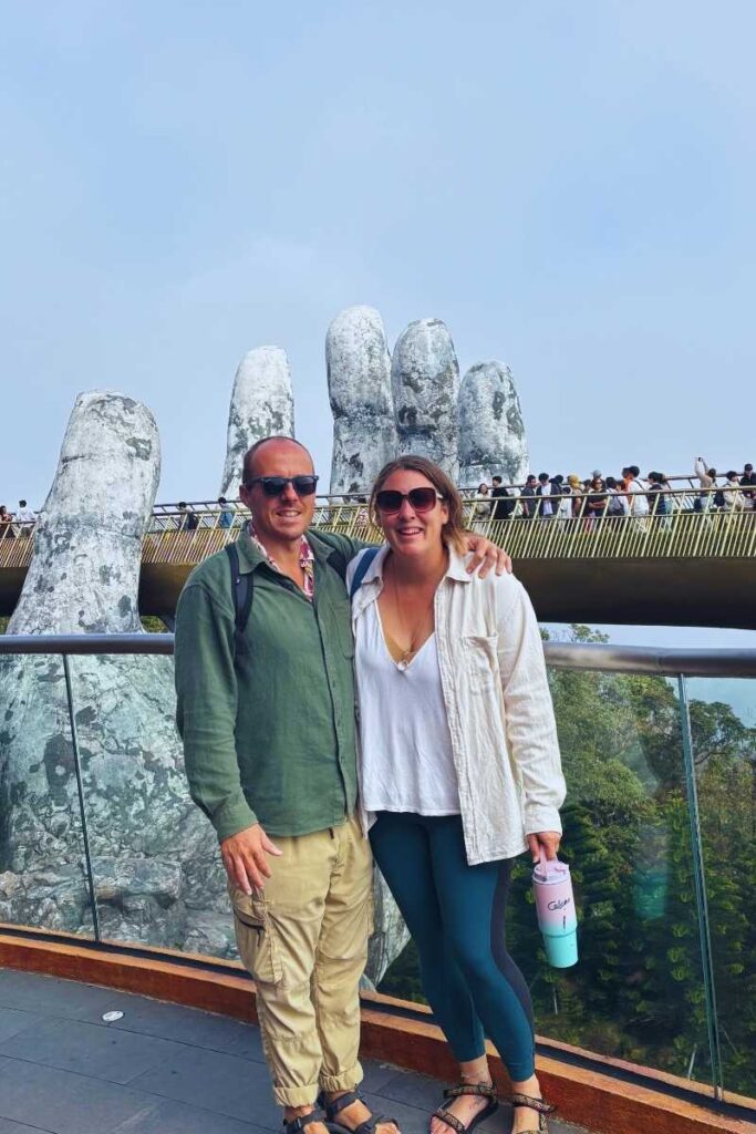 Two visitors posing in front of the Golden Bridge at Ba Na Hills, with large stone hand sculptures and a crowd of people walking across the bridge in the background.