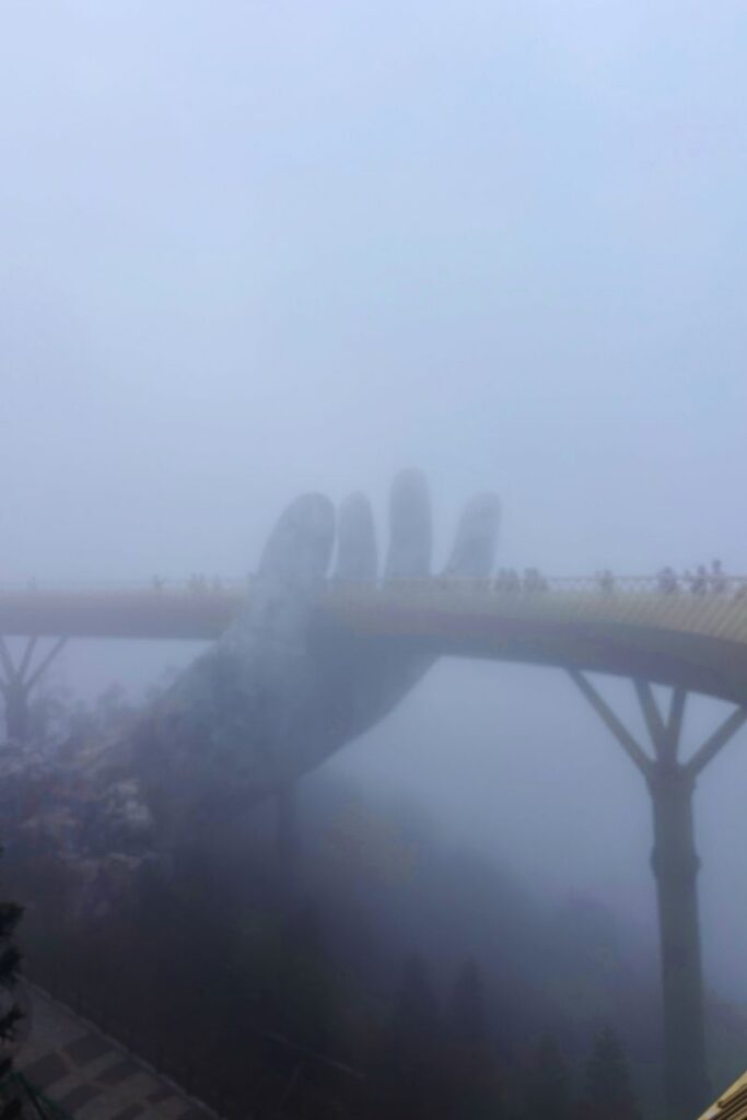 Golden Bridge at Ba Na Hills partially obscured by heavy fog, with the giant stone hand sculpture holding the walkway and visitors walking across.