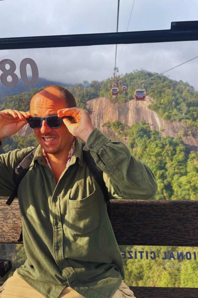 Man adjusting sunglasses in front of the Ba Na Hills cable car line, with several gondolas traveling over a forested mountain slope and large rock formations.