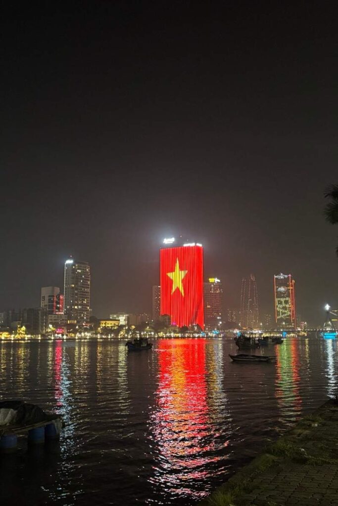 Night view of Da Nang’s skyline from the Han River, with boats on the water and a brightly lit building displaying the Vietnamese flag.