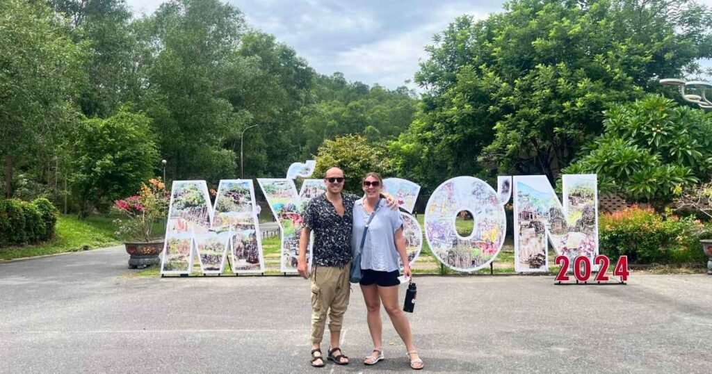 Two smiling travelers pose in front of the large “Mỹ Sơn 2024” entrance sign, surrounded by lush greenery and jungle-lined roads at My Son Sanctuary in Vietnam.