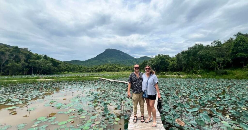 Two smiling travelers stand on a narrow boardwalk over a lotus pond, with Cat’s Tooth Mountain (Răng Mèo) towering in the distance behind them.