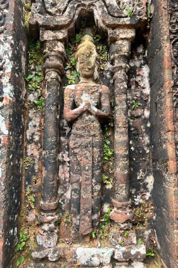 An ancient stone carving of a standing deity in a praying posture, nestled between two pillars and surrounded by moss and vegetation growing from the brick temple wall at My Son Sanctuary.