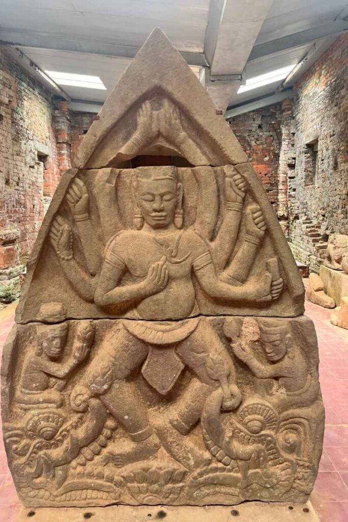 A detailed relief carving of a multi-armed Hindu deity in a dynamic pose, framed by an arched temple motif, housed within the museum area at My Son Sanctuary.