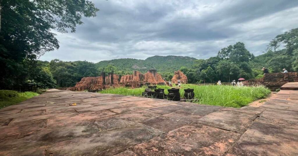 A stone pathway leads toward the central temple group at My Son Sanctuary, where tourists explore the grassy, ruin-filled valley surrounded by dense forest.