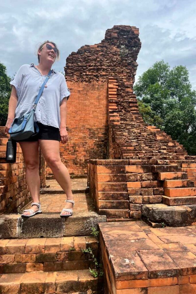 A woman stands smiling atop ancient red-brick steps at My Son Sanctuary with the remains of a partially crumbled temple behind her.