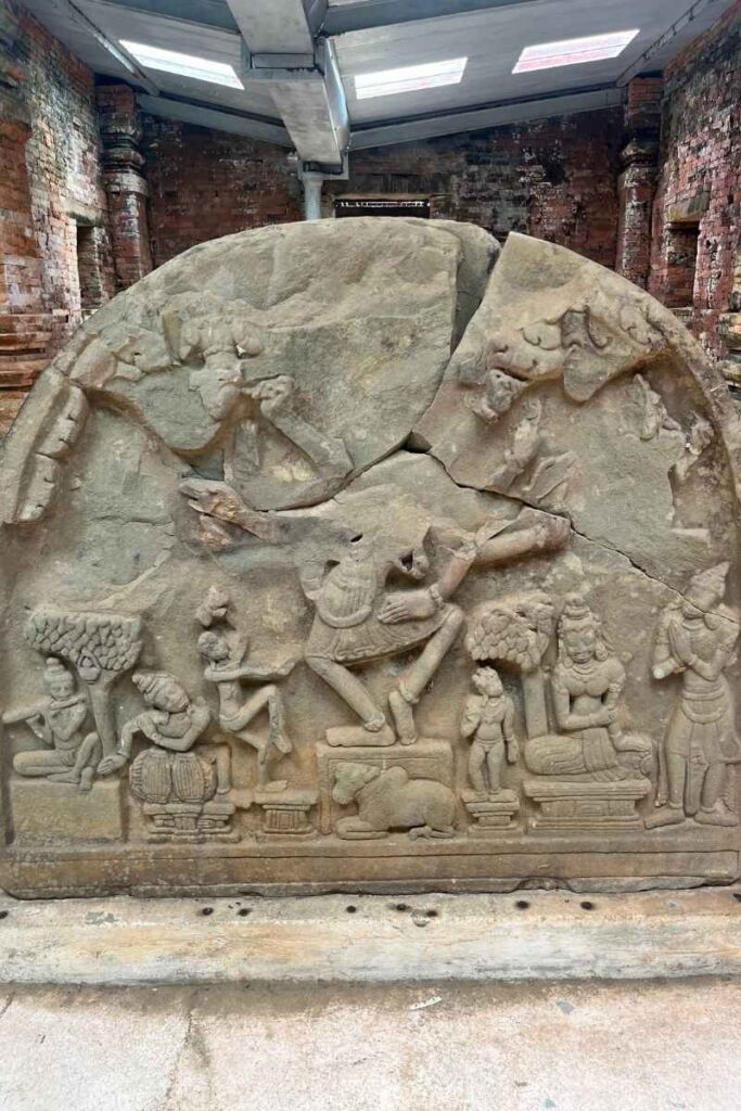 A large, semi-circular stone carving featuring a detailed relief of a mythological scene with musicians, dancers, and seated deities, partially cracked and weathered with age, housed in a museum setting at My Son Sanctuary.