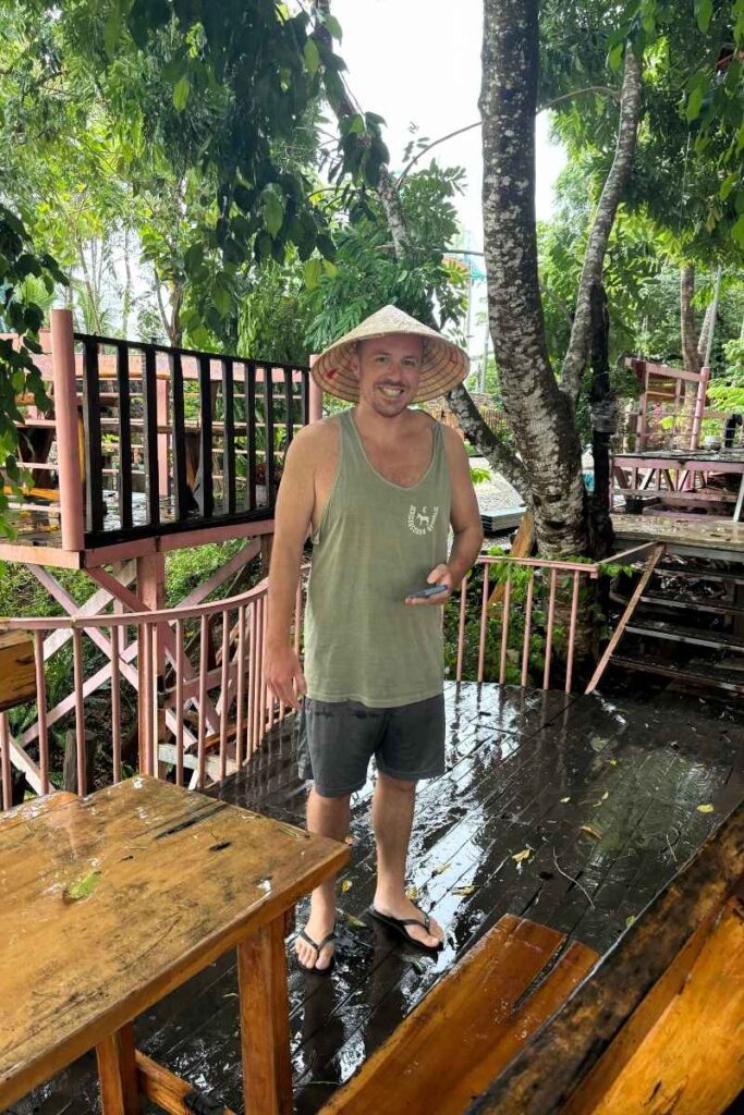 Man wearing a nón lá hat and holding a phone on the wooden deck of a rustic lodge near Cát Tiên accommodation, surrounded by lush greenery after rain.