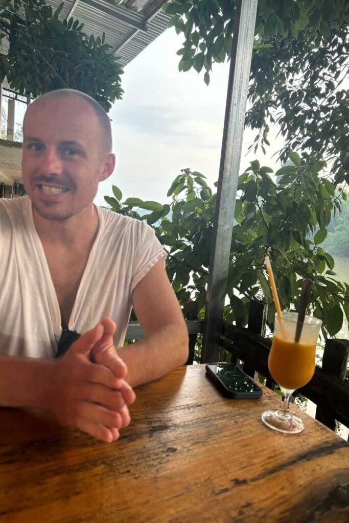 Man smiling at an outdoor café table near Cát Tiên National Park, with leafy trees and drinks in the background.