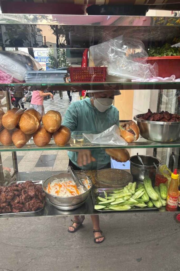 Street food vendor assembling fresh bánh mì sandwiches behind a glass case filled with pickled vegetables, cucumbers, grilled meat, and baguettes in Ho Chi Minh City.