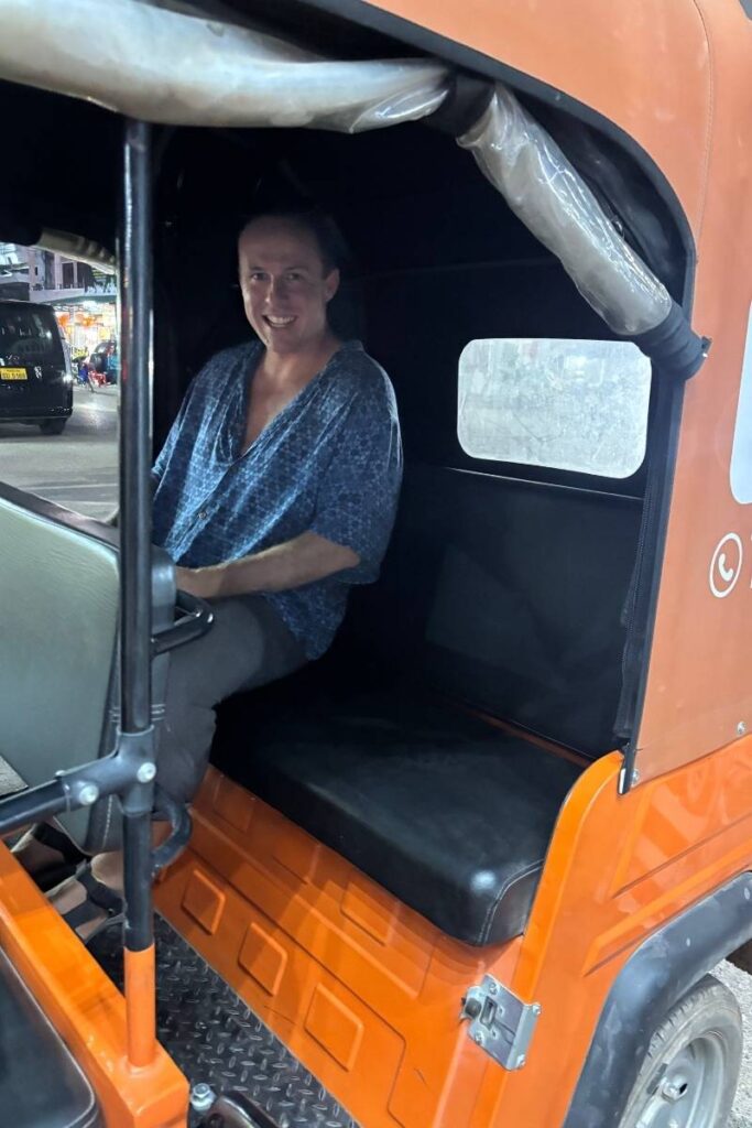 Person smiling while seated in the back of a bright orange KokKok, a local tuk-tuk-style rideshare vehicle in Laos. The scene captures nighttime city traffic in the background, illustrating a common and convenient alternative to the Grab or Uber app in Southeast Asia.
