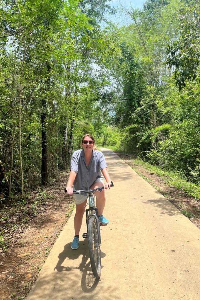 Woman biking along a sunny path through dense jungle in national park Cát Tiên, surrounded by vibrant greenery and tall trees.
